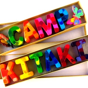 May include: Two boxes containing colorful crayon letters spelling out "CAMP KITAKI". The boxes are made of cardboard and have a natural brown color. The letters are made of crayon wax and have a rainbow color scheme. The top box has a small, colorful butterfly decoration. The bottom box has a small, colorful leaf decoration.