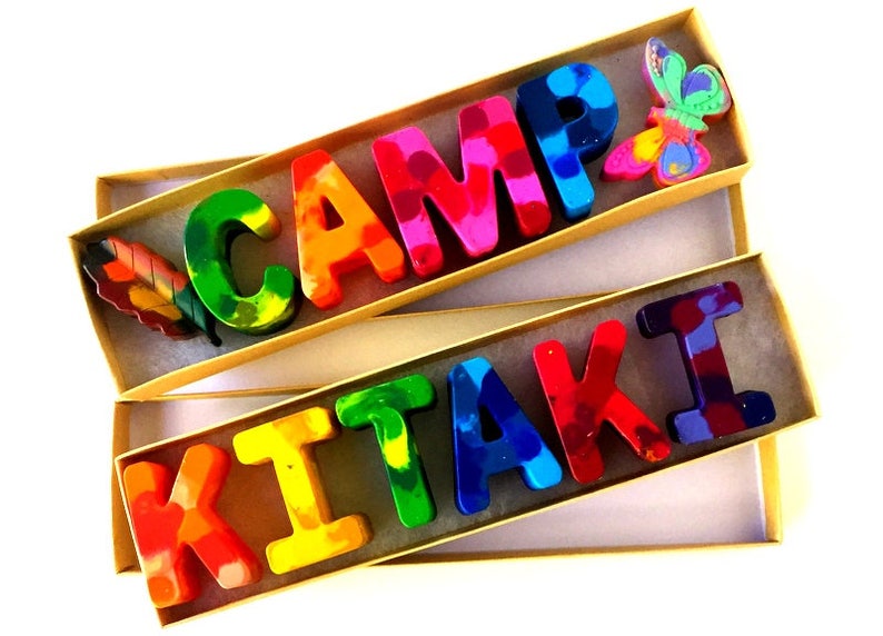 May include: Two boxes containing colorful crayon letters spelling out "CAMP KITAKI". The boxes are made of cardboard and have a natural brown color. The letters are made of crayon wax and have a rainbow color scheme. The top box has a small, colorful butterfly decoration. The bottom box has a small, colorful leaf decoration.
