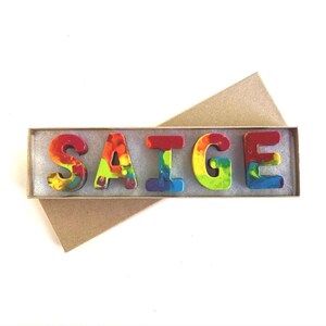 May include: A set of colorful crayon letters spelling the name "SAIGE" in a brown cardboard box.
