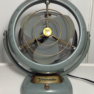 Puede incluir: Un ventilador oscilante Vornado vintage gris con una jaula de ventilador de metal dorado y un interruptor de metal dorado. El ventilador tiene una base redonda y un brazo curvo.