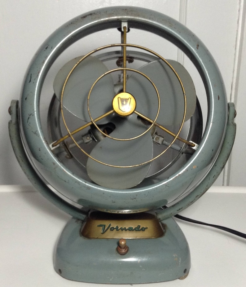 Puede incluir: Un ventilador oscilante Vornado vintage gris con una jaula de ventilador de metal dorado y un interruptor de metal dorado. El ventilador tiene una base redonda y un brazo curvo.