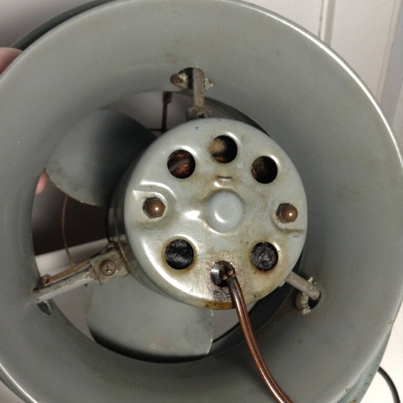 Puede incluir: Primer plano de la parte posterior de un ventilador de metal gris con un cable de cobre conectado. El ventilador tiene una carcasa met&aacute;lica circular con seis agujeros alrededor del centro. Las aspas del ventilador son visibles en el fondo.