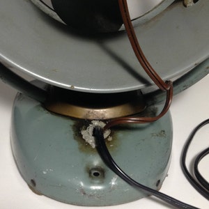 Puede incluir: Un ventilador vintage de metal azul claro con un cable marr&oacute;n. El ventilador est&aacute; en posici&oacute;n inclinada, mostrando la parte inferior de la base. La base tiene un anillo de lat&oacute;n alrededor de la parte superior y un cable negro que sale de la parte inferior.