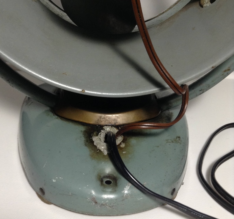 Puede incluir: Un ventilador vintage de metal azul claro con un cable marr&oacute;n. El ventilador est&aacute; en posici&oacute;n inclinada, mostrando la parte inferior de la base. La base tiene un anillo de lat&oacute;n alrededor de la parte superior y un cable negro que sale de la parte inferior.