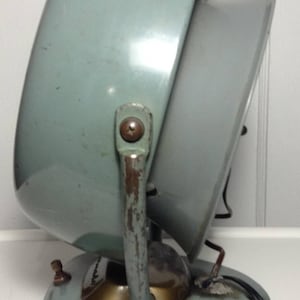 Puede incluir: Un calentador el&eacute;ctrico vintage de color azul claro con un asa de metal y un ventilador redondo inclinado. El calentador tiene una base dorada y un cable.