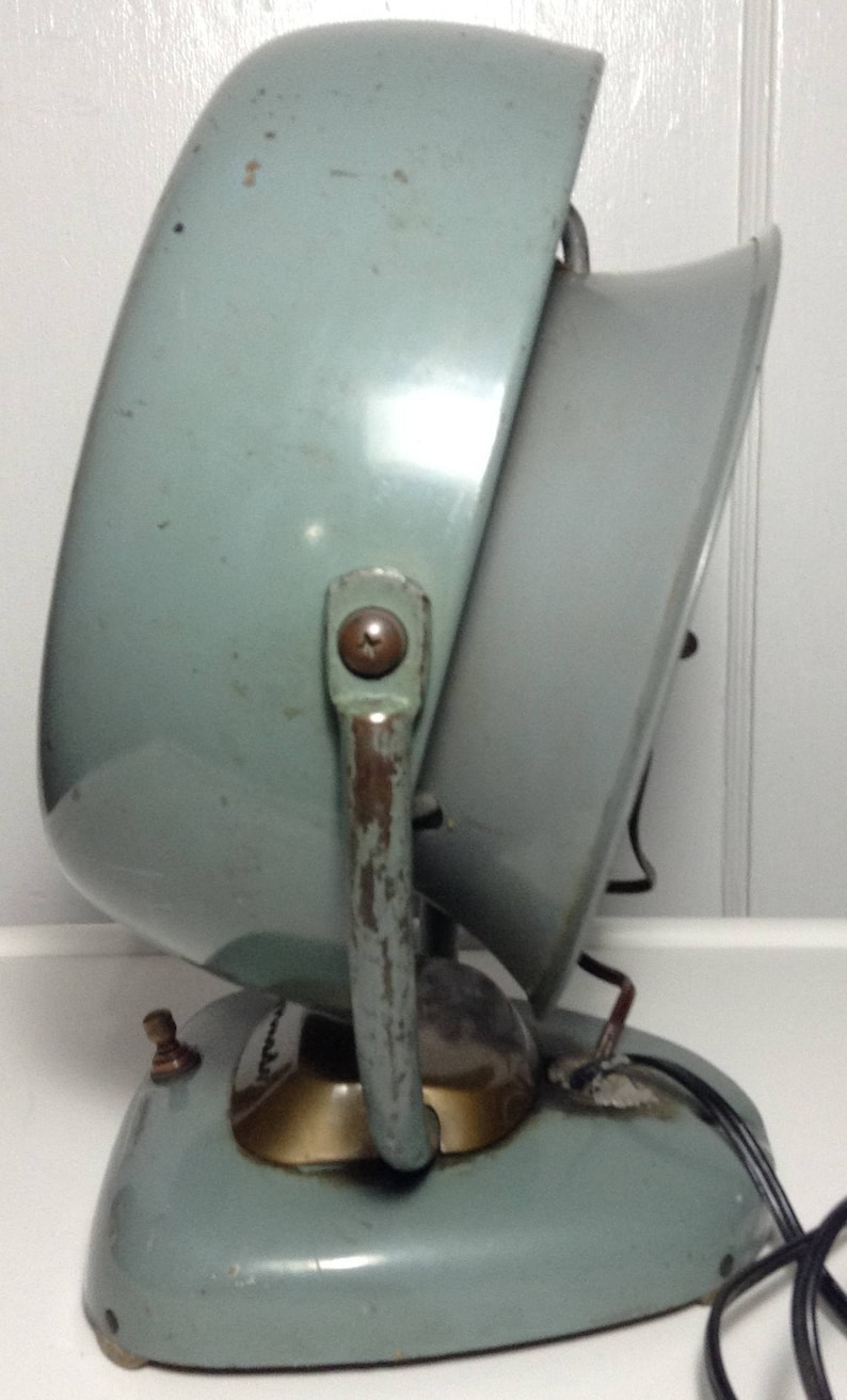 Puede incluir: Un calentador el&eacute;ctrico vintage de color azul claro con un asa de metal y un ventilador redondo inclinado. El calentador tiene una base dorada y un cable.