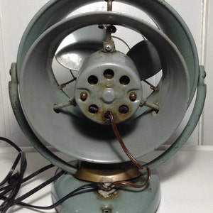 Puede incluir: Un ventilador oscilante vintage gris con una base circular y una rejilla de metal. El ventilador tiene un cable de alimentaci&oacute;n marr&oacute;n y un motor visible con seis agujeros.