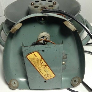 Puede incluir: Un ventilador Vornado vintage con un cuerpo de metal azul claro y un cable negro. El ventilador tiene una etiqueta en la parte inferior que dice "Vornado Model D16C1 Serial No. 050 278141 UL".