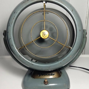 Puede incluir: Un ventilador Vornado vintage azul y dorado con una jaula met&aacute;lica circular y un solo interruptor en la base.