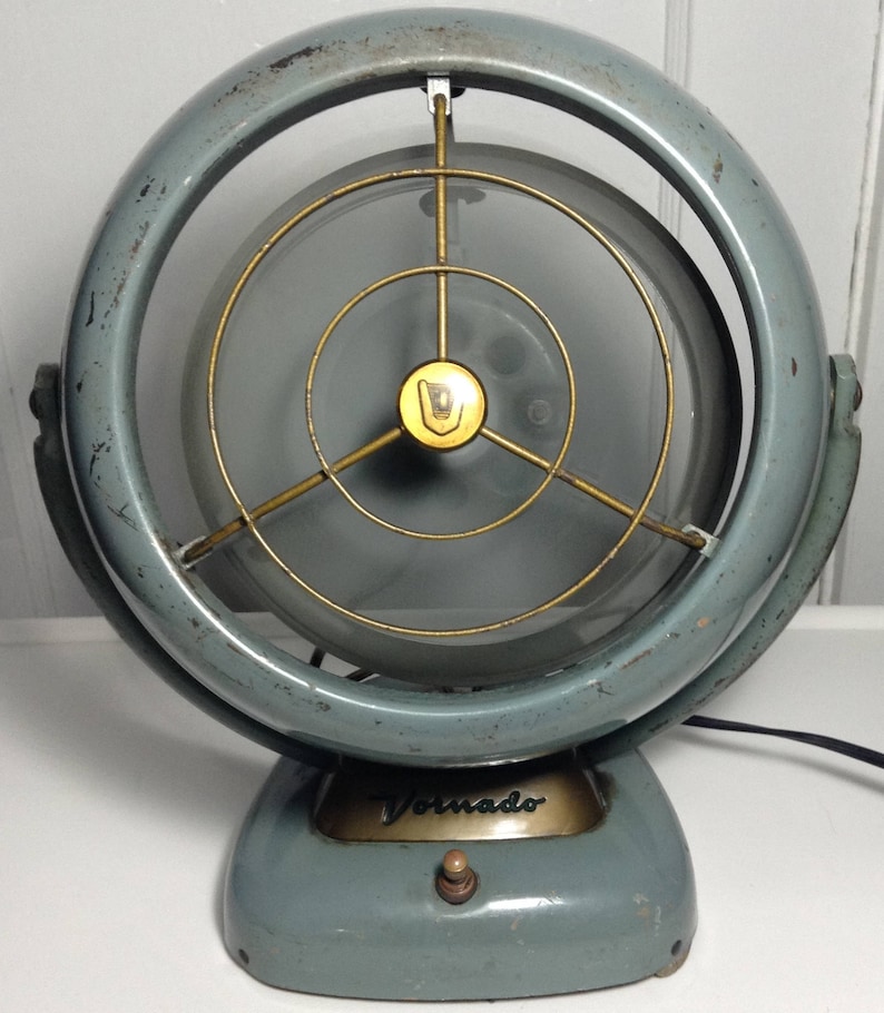 Puede incluir: Un ventilador Vornado vintage azul y dorado con una jaula met&aacute;lica circular y un solo interruptor en la base.