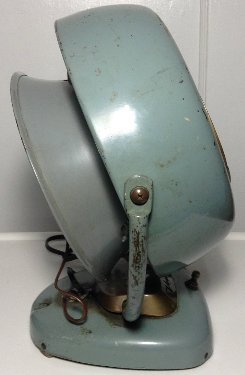 Puede incluir: Un calentador el&eacute;ctrico vintage de color azul claro con un asa de metal y una base redonda. El calentador tiene una rejilla de metal y un cable.