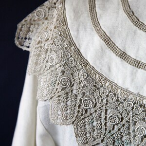 May include: A white linen shirt with a delicate lace collar. The lace is a light brown color and has a floral pattern.