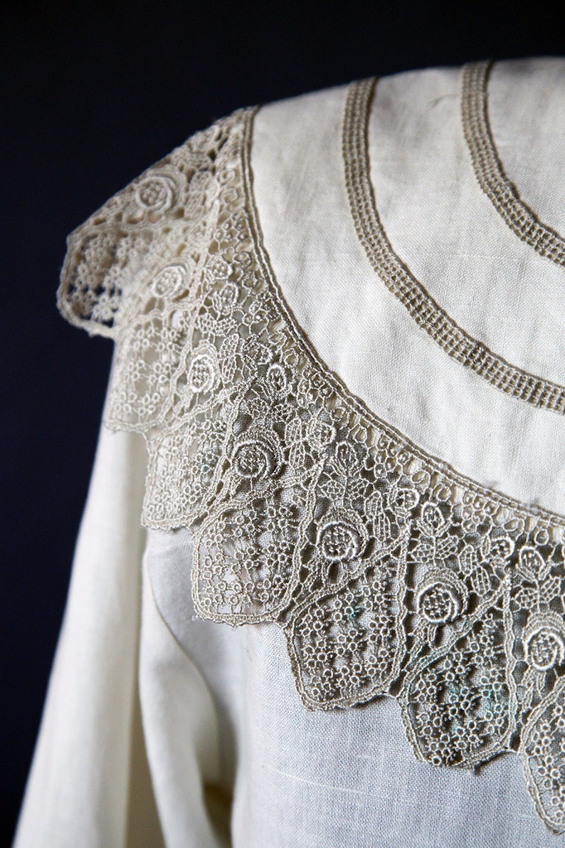 May include: A white linen shirt with a delicate lace collar. The lace is a light brown color and has a floral pattern.