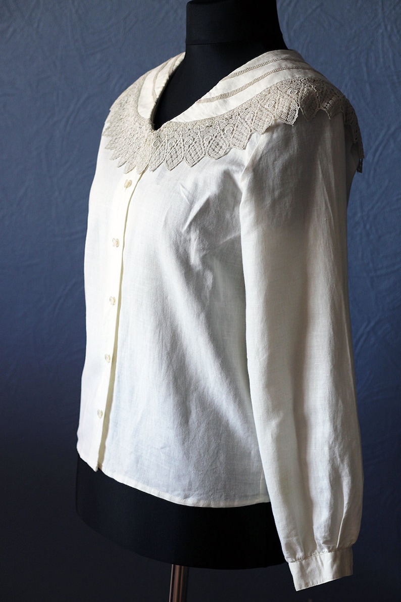May include: A white long-sleeved button-up shirt with a large, lacy collar. The collar is white with a delicate, intricate design.