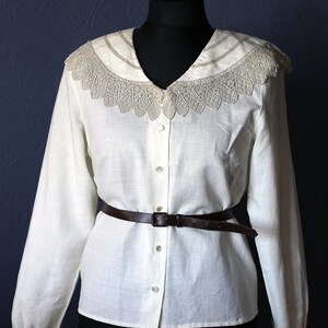 May include: A white linen blouse with a lace collar and a brown leather belt. The blouse has a button-up front and long sleeves.