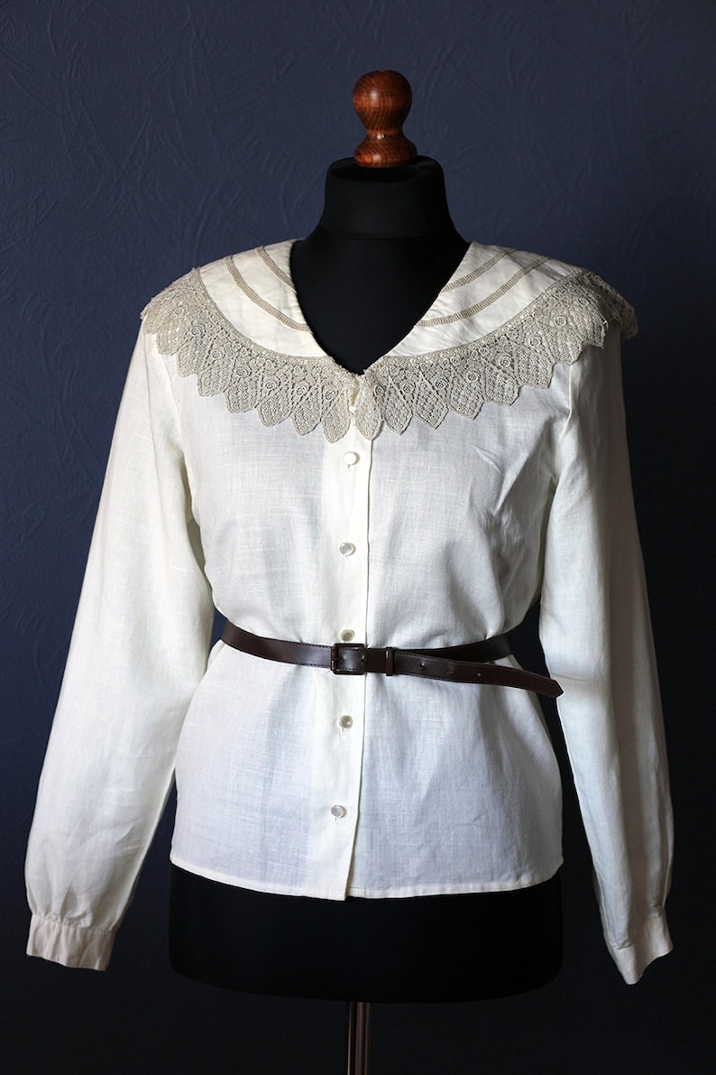 May include: A white linen blouse with a lace collar and a brown leather belt. The blouse has a button-up front and long sleeves.