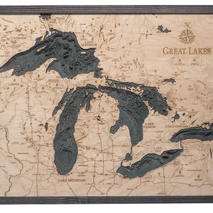 May include: A framed wooden bathymetric map of the Great Lakes featuring laser-cut layers of light-colored wood for the land and dark gray recessed layers for the water depths. The map includes engraved labels for the lakes and surrounding states.