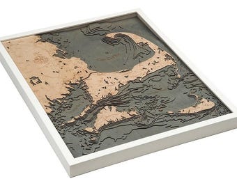 May include: A three-dimensional wooden bathymetric map of Cape Cod framed in white. The map features layered light wood landmasses against a dark gray water background, with a compass rose and engraved text labels visible on the surface.