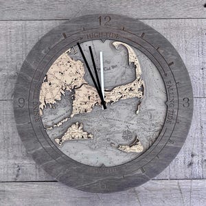 May include: A round wooden wall clock featuring a detailed bathymetric map of Cape Cod, Martha's Vineyard, and Nantucket. The dark gray frame displays the numbers 3, 6, 9, 12 and text indicating tide levels: High Tide, Falling Tide, Low Tide, Rising Tide.