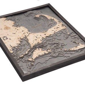 May include: A framed three-dimensional bathymetric wood map of Cape Cod, Massachusetts. The map features light-colored wooden landmasses set against dark gray water layers with engraved depth contours, a compass rose, and labeled geographic locations.