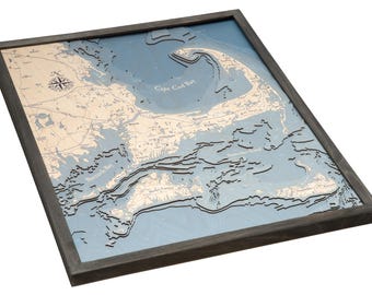 May include: A framed 3D bathymetric wood map of Cape Cod Bay and surrounding coastal areas. The map features layered blue water depths and light tan landmasses with black text labels, all enclosed in a dark gray wooden frame.