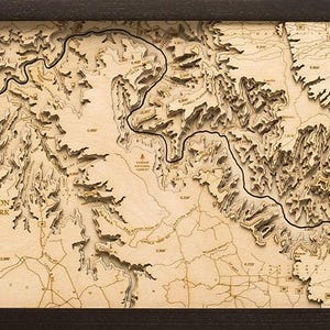 May include: A framed, three-dimensional wooden topographic map of the Grand Canyon National Park. The light-colored wood features laser-cut layers showing the river path, canyon ridges, and elevation markers, all enclosed in a dark brown frame.