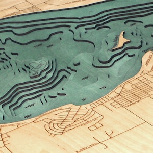 K&ouml;nnte beinhalten: Eine Holzkarte eines Sees mit Tiefenkonturen, die in Fu&szlig; angegeben sind. Der See ist in blaugr&uuml;ner Farbe gehalten und das umliegende Land ist hellbraun. Die Karte ist aus Holz gefertigt und hat einen 3D-Effekt.