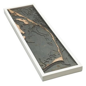 May include: A framed wooden map of the Outer Banks, North Carolina. The map is in shades of gray and brown, with the coastline highlighted in brown. The map is framed in a white frame.