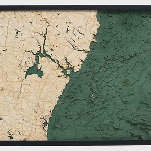 May include: A framed 3D bathymetric wood map featuring a light tan landmass with detailed black road and town labels alongside a deep green textured ocean area with carved depth contours and a small compass rose etched into the water surface.