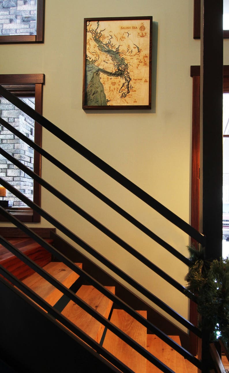 K&ouml;nnte beinhalten: Eine Holzkarte der Salish Sea h&auml;ngt an einer Wand mit einem Holzrahmen. Die Karte ist hellbraun mit blauen Wassermerkmalen. Die Karte h&auml;ngt an einer Wand mit einer Holztreppe im Vordergrund.
