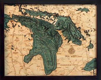 May include: A framed 3D wood bathymetric map of Lake Huron and surrounding land areas. The water is depicted in layered shades of teal and blue with depth contours, while the land is light wood with black text labels and a compass rose.