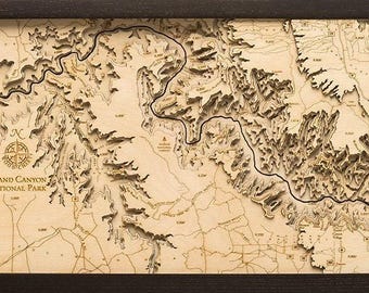 May include: A rectangular framed 3D wooden topographic map of Grand Canyon National Park. The light wood surface features laser-cut elevation layers, a winding river path, a compass rose, and the text Grand Canyon National Park.