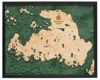 May include: A framed 3D wooden bathymetric map of Drummond Island featuring light wood landmasses against deep green water layers. The map includes a compass rose and the text Drummond Island laser-etched into the surface.