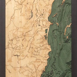 May include: A vertical wooden bathymetric map of Belize featuring laser-cut land topography in light natural wood and deep green layered water depths. The map includes a compass rose, labeled cities like Belize City and Belmopan, and a black frame.