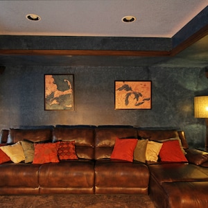 K&ouml;nnte beinhalten: Ein braunes Ledersofa in L-Form mit Kissen in Rot-, Gr&uuml;n- und Braunt&ouml;nen. Das Sofa steht in einem Wohnzimmer mit einer Lampe und zwei gerahmten Karten an der Wand.