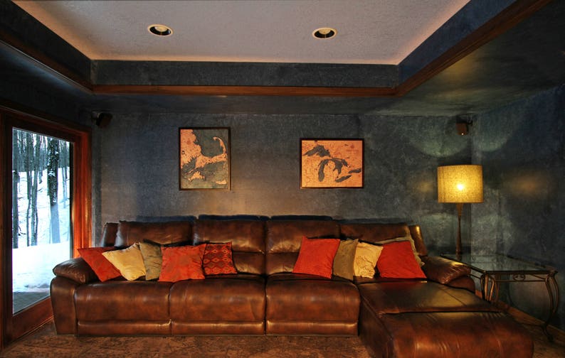 K&ouml;nnte beinhalten: Ein braunes Ledersofa in L-Form mit Kissen in Rot-, Gr&uuml;n- und Braunt&ouml;nen. Das Sofa steht in einem Wohnzimmer mit einer Lampe und zwei gerahmten Karten an der Wand.