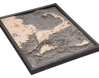 May include: A three-dimensional wooden bathymetric map of Cape Cod, Massachusetts, framed in dark wood. The map features light-colored landmasses against a dark gray water surface with layered depth contours and a compass rose.