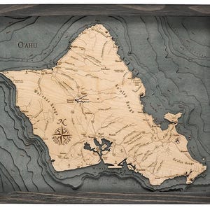 May include: A framed 3D wood chart of the island of O'ahu featuring a light-colored wooden landmass with engraved text, roads, and landmarks, set against layered, dark gray, recessed bathymetric contours of the surrounding ocean.