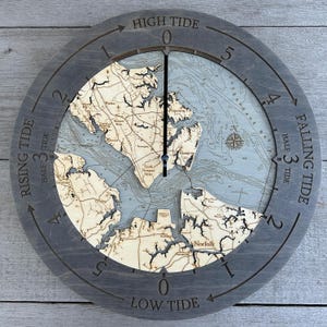 May include: A round, gray tide clock with a detailed map of a coastal area. The clock face features a light blue and beige map, with the words "HIGH TIDE" and "LOW TIDE" marked. The clock hands point to the current tide level.