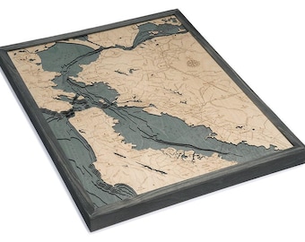 May include: A rectangular wooden bathymetric map of the San Francisco Bay area featuring layered light tan landmasses and deep blue water depths. The map is encased in a dark gray wooden frame and includes a compass rose and city labels.