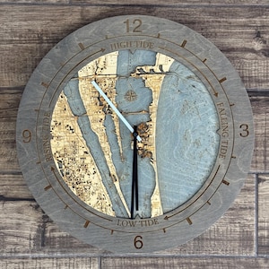 May include: A circular wooden wall clock featuring a laser-cut nautical map of a coastal area. The grey frame displays the numbers 3, 6, 9, and 12, with text indicating High Tide, Falling Tide, Low Tide, and Rising Tide. It has black and white hands.