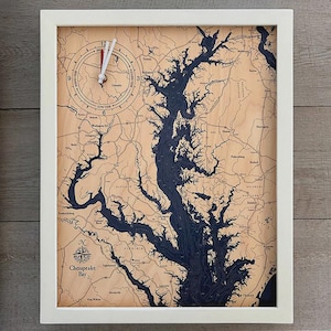 May include: A framed wall clock featuring a detailed map of Chesapeake Bay. The map is in shades of blue and brown, with a clock face at the top. The clock has white hands and a white frame.
