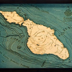 May include: A framed 3D wood chart of Catalina, California, featuring a light-colored wooden island map set against layered blue water depths. The map includes text labels for locations like Avalon and Two Harbors, bathymetric depth markers, and a compass rose.