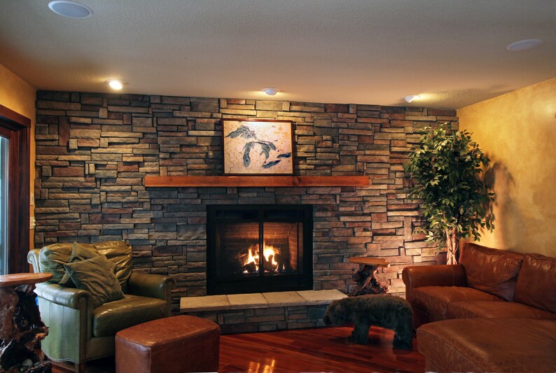 K&ouml;nnte beinhalten: Ein Wohnzimmer mit einem Steinkamin und einem gro&szlig;en braunen Ledersofa. Der Kamin hat einen schwarzen Metallrost und ein Feuer brennt darin. Vor dem Kamin steht ein gr&uuml;ner Sessel und ein kleiner Holztisch mit einer Pflanze darauf.