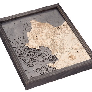 May include: A framed wooden map of a coastal area with detailed topography and depth markings. The map is in shades of brown and gray, with a compass rose in the lower right corner. The map is framed in a dark gray wood frame.