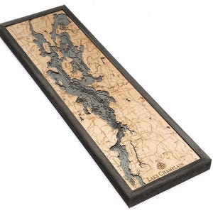 May include: A rectangular wooden wall map of Lake Champlain featuring a layered, three-dimensional bathymetric design. The map displays light tan land areas with engraved topographical details and dark gray water depths, all enclosed in a dark frame.