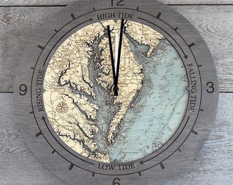 May include: A round wooden wall clock featuring a nautical map of the Chesapeake Bay. The face displays land in tan and water in light blue, with black clock hands. The frame includes the text High Tide, Falling Tide, Low Tide, and Rising Tide.