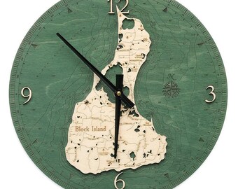 Op de afbeelding: Een ronde wandklok met een licht houten kaart van Block Island in het midden op een groene achtergrond met bathymetrische hoogtelijnen. De klok heeft zwarte wijzers, grote cijfers op 12, 3, 6 en 9, en een kompasroos aan de rechterkant.