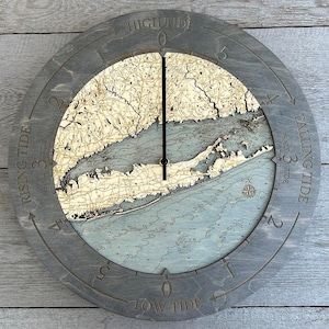 May include: A wooden clock with a map of Long Island, New York, on the face. The clock has a gray wooden frame and a black hand. The clock face is labeled with "High Tide", "Low Tide", "Rising Tide", and "Falling Tide".