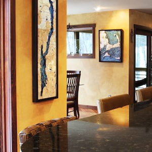 K&ouml;nnte beinhalten: Eine schwarze Granit-Arbeitsplatte mit einem geflochtenen Barhocker aus Weide davor. Die Wand hinter der Arbeitsplatte ist in einem warmen Gelbton gehalten und zeigt zwei gerahmte Holz-Karten von Seen.